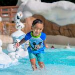A young child plays in the pool at Disney's Blizzard Beach Water Park. Olaf from Frozen is in the background, along with several Snowgies.
