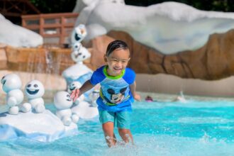 A young child plays in the pool at Disney's Blizzard Beach Water Park. Olaf from Frozen is in the background, along with several Snowgies.