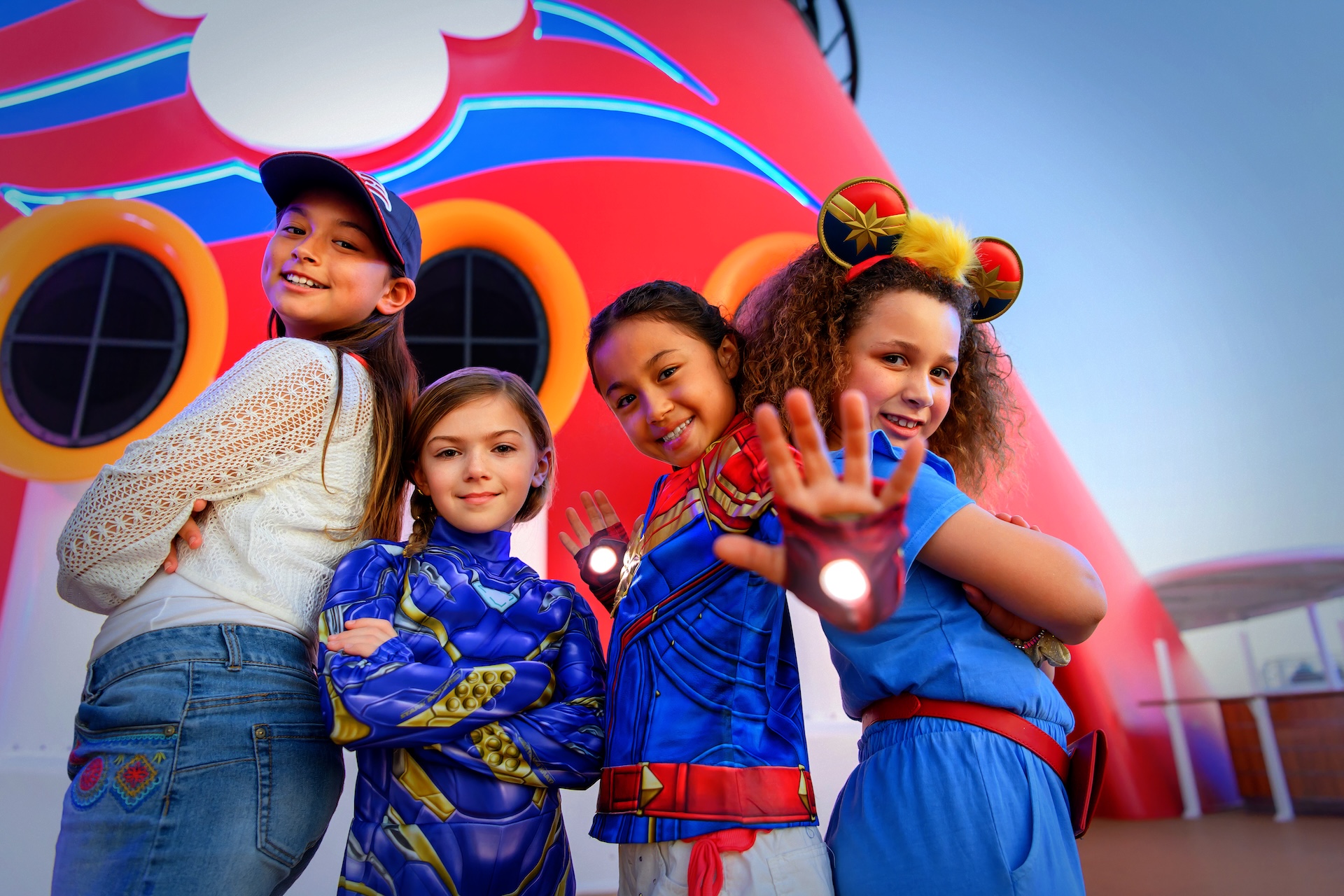 Four children pose in superhero themed outfits on a Disney Cruise Line deck in front of the ship funnel