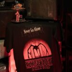 Stranger Things 5 Halloween Horror Nights merch: a Never Go Alone T-shirt with Vecna and Demogorgon acrylic figures on the Infernal Carnival display