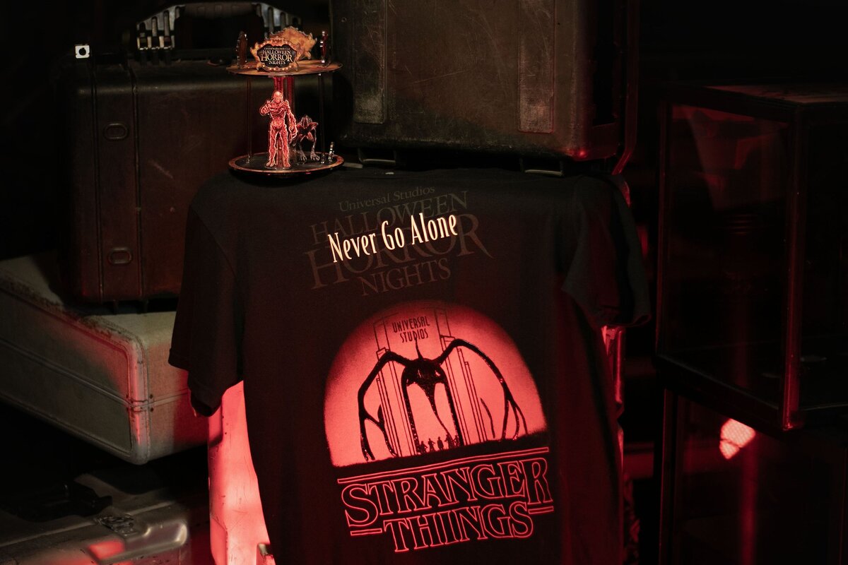 Stranger Things 5 Halloween Horror Nights merch: a Never Go Alone T-shirt with Vecna and Demogorgon acrylic figures on the Infernal Carnival display