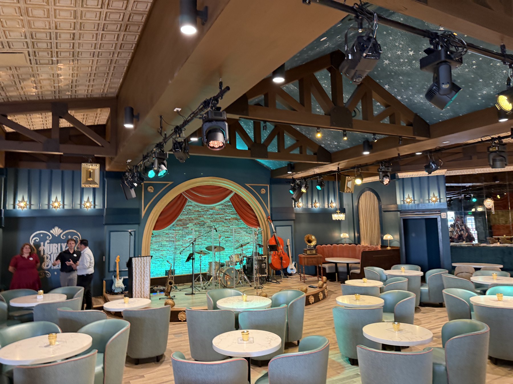Blue lounge chairs, round tables and an arched stage inside Hurly-Burly at Disney’s BoardWalk.