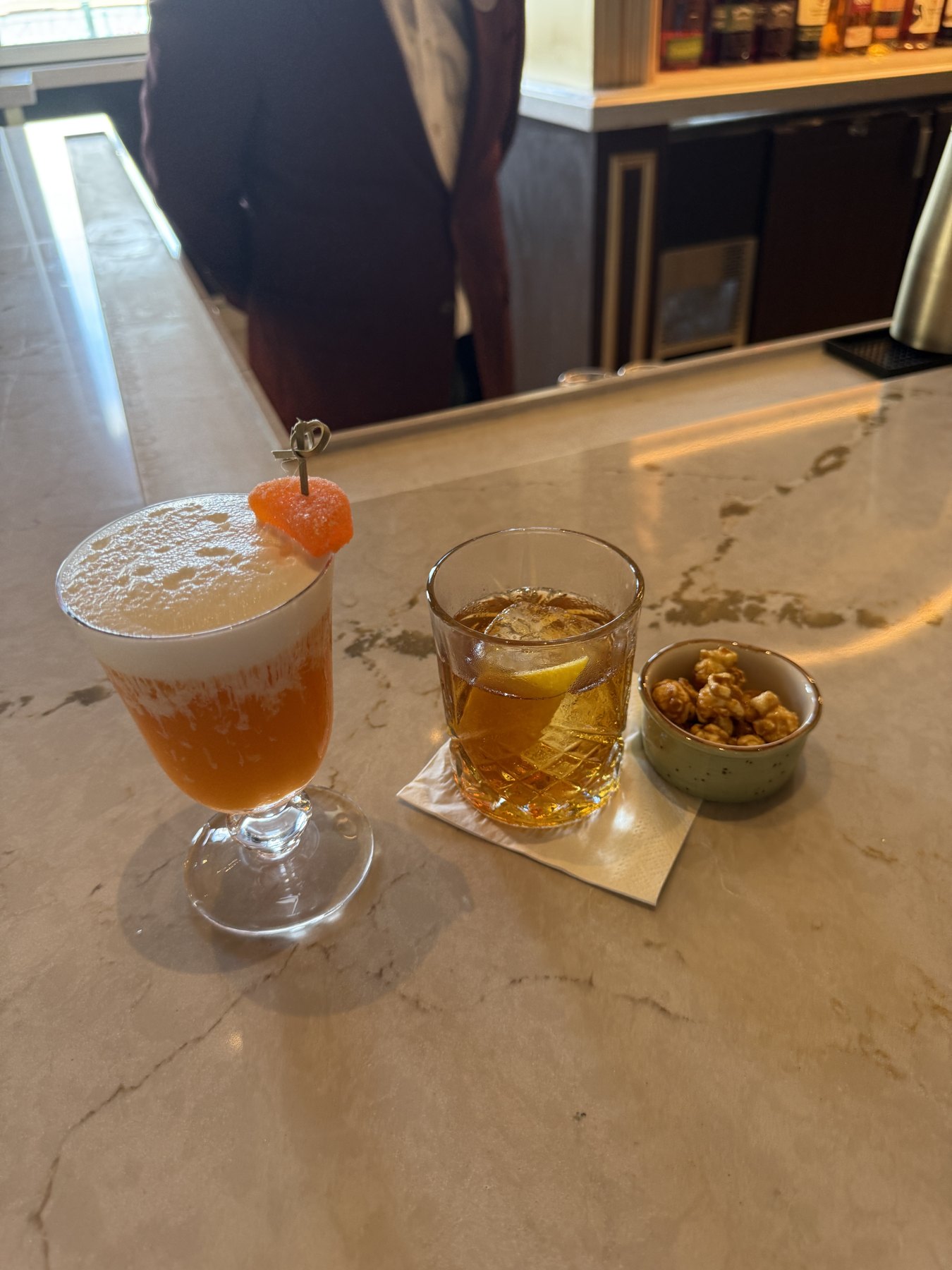 An Old Fashioned in a rocks glass and a foamy orange drink in a stemmed glass on the bar.