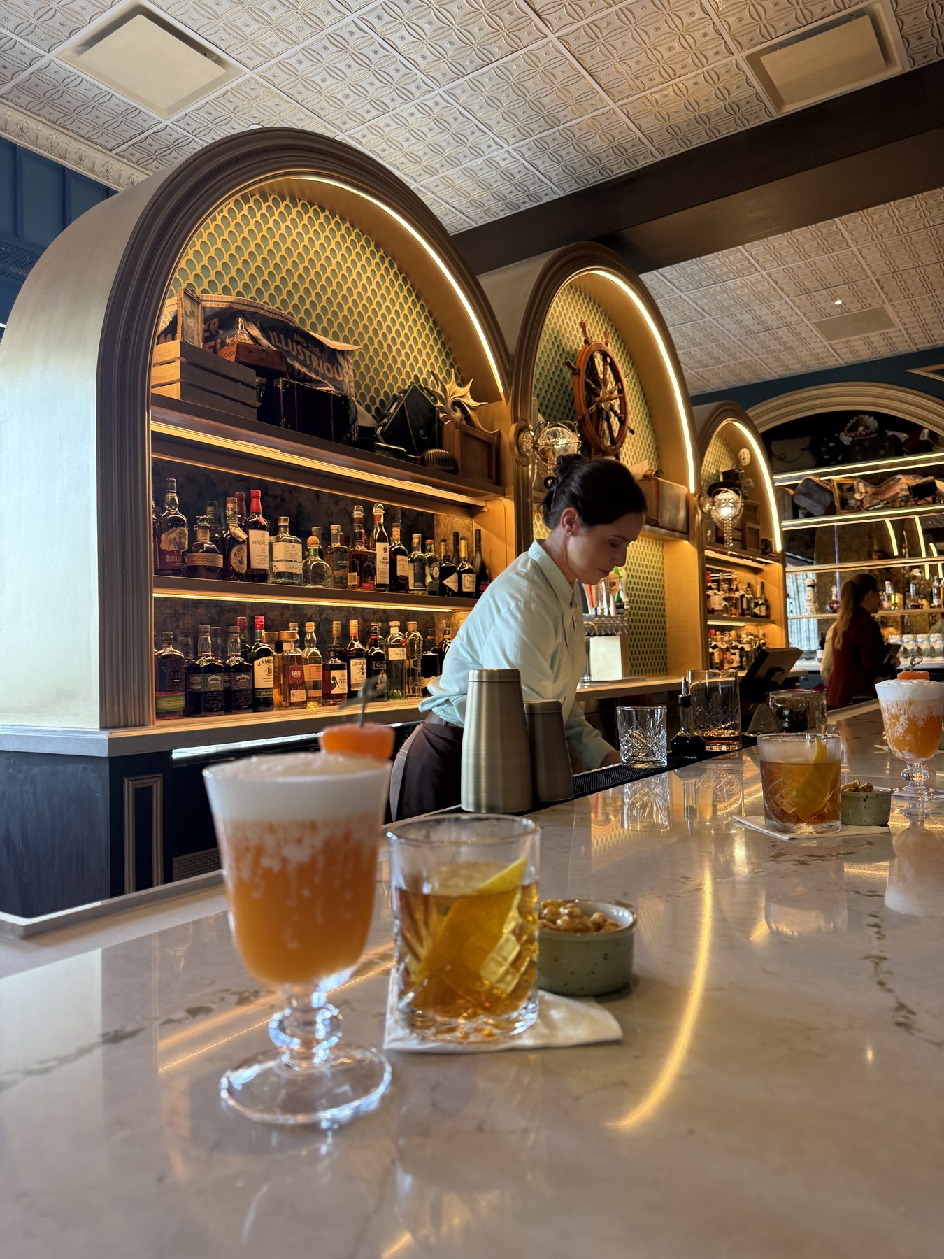 Two drinks in the foreground on the pale countertop beneath Hurly-Burly’s illuminated bar shelves.