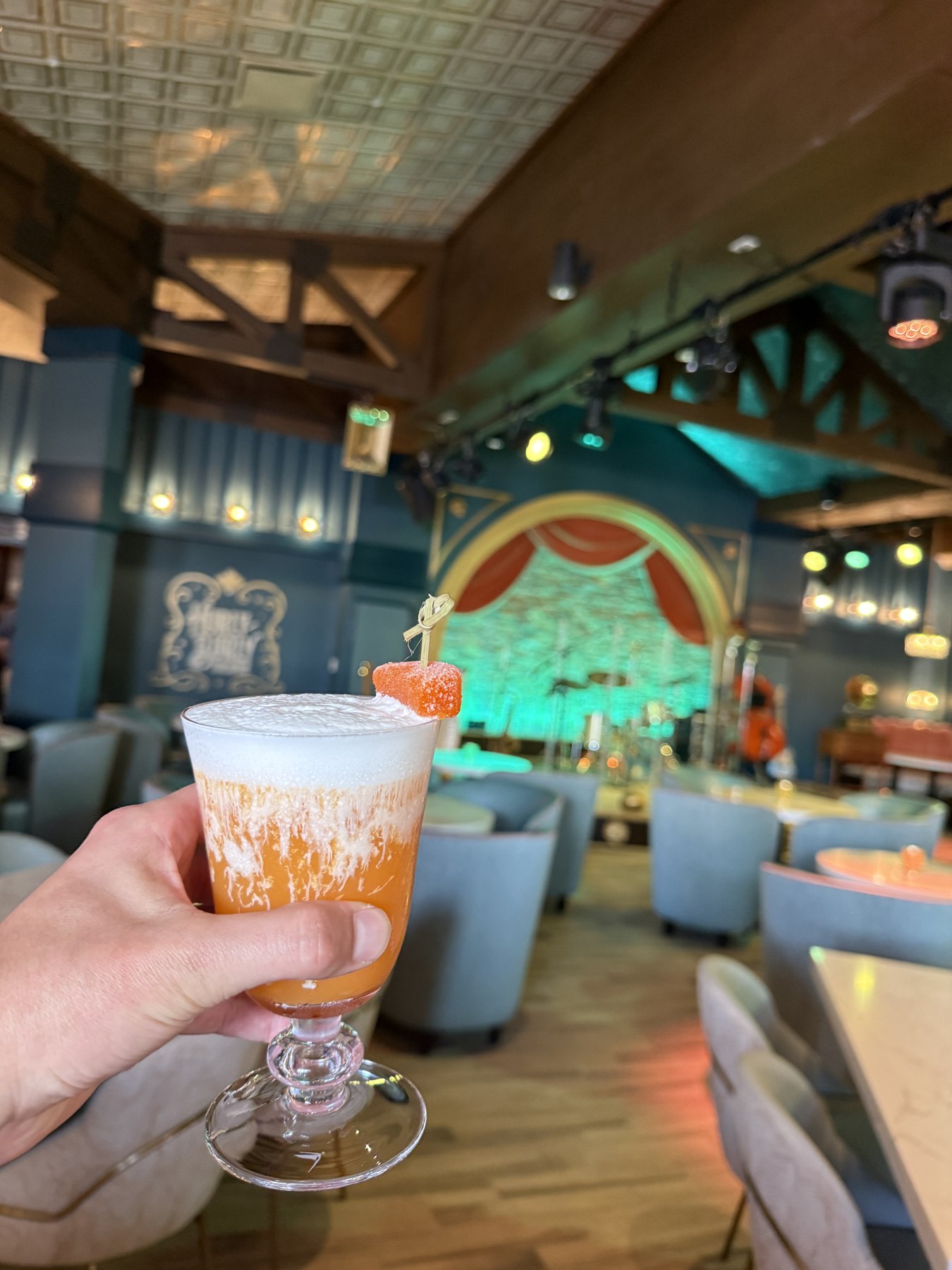 A hand holds a foamy orange drink with Hurly-Burly’s stage and seating area in the background.