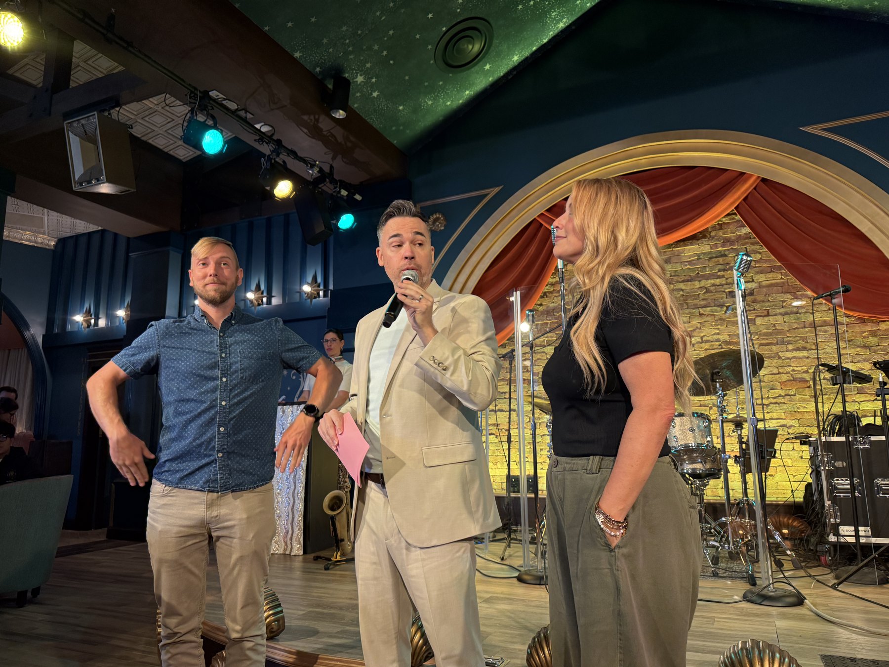 Three adults stand on Hurly-Burly’s stage while the center presenter speaks into a microphone.