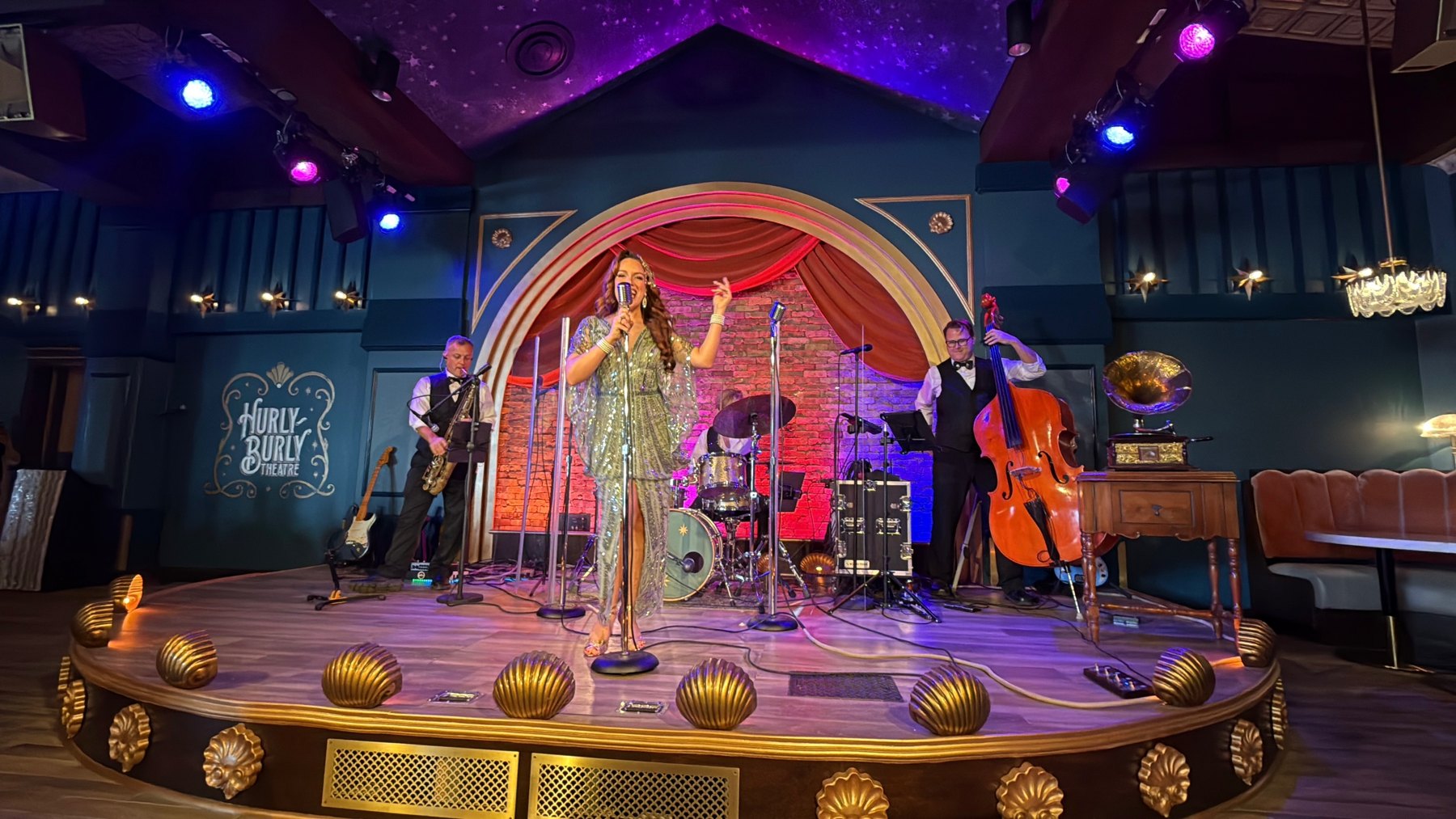 A vocalist in a silver outfit performs with a saxophonist, upright bassist and drummer on Hurly-Burly’s stage.