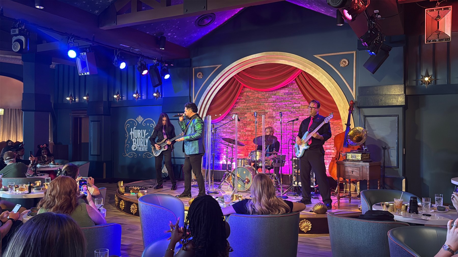 A singer, guitarist, bassist and drummer perform on Hurly-Burly’s stage, with guests seated nearby.