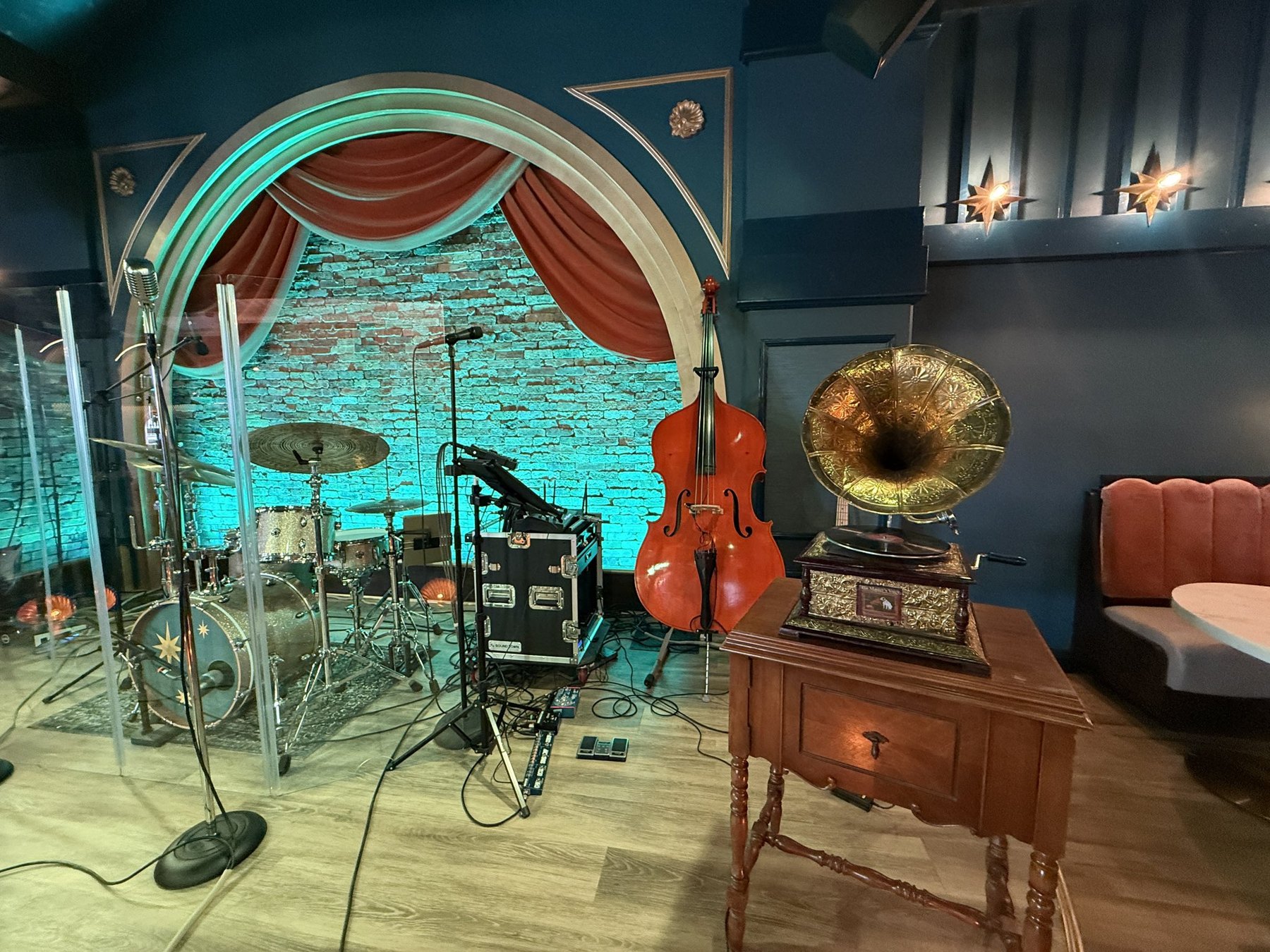 A curved stage with drums, upright bass, coral-colored curtains and a vintage-style gramophone nearby.